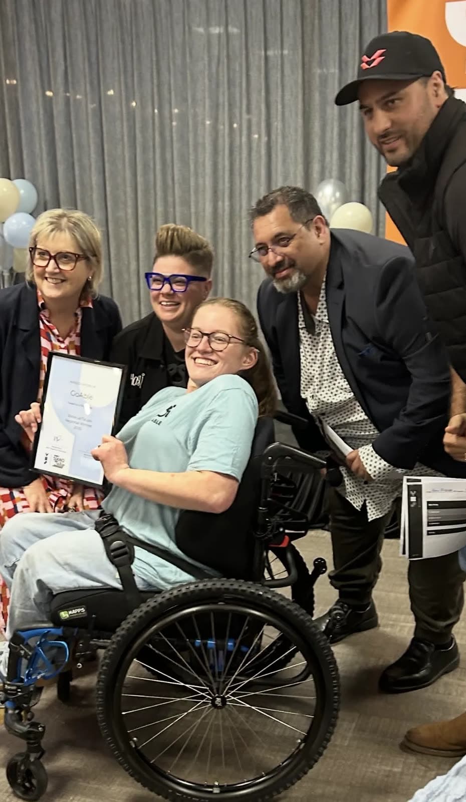 Daisy Hoe holding a Go Able regional winner certificate, surrounded by supporters