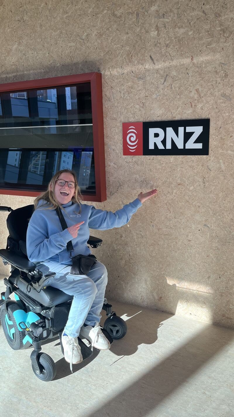 Daisy Hoe smiling in her wheelchair in front of the RNZ logo