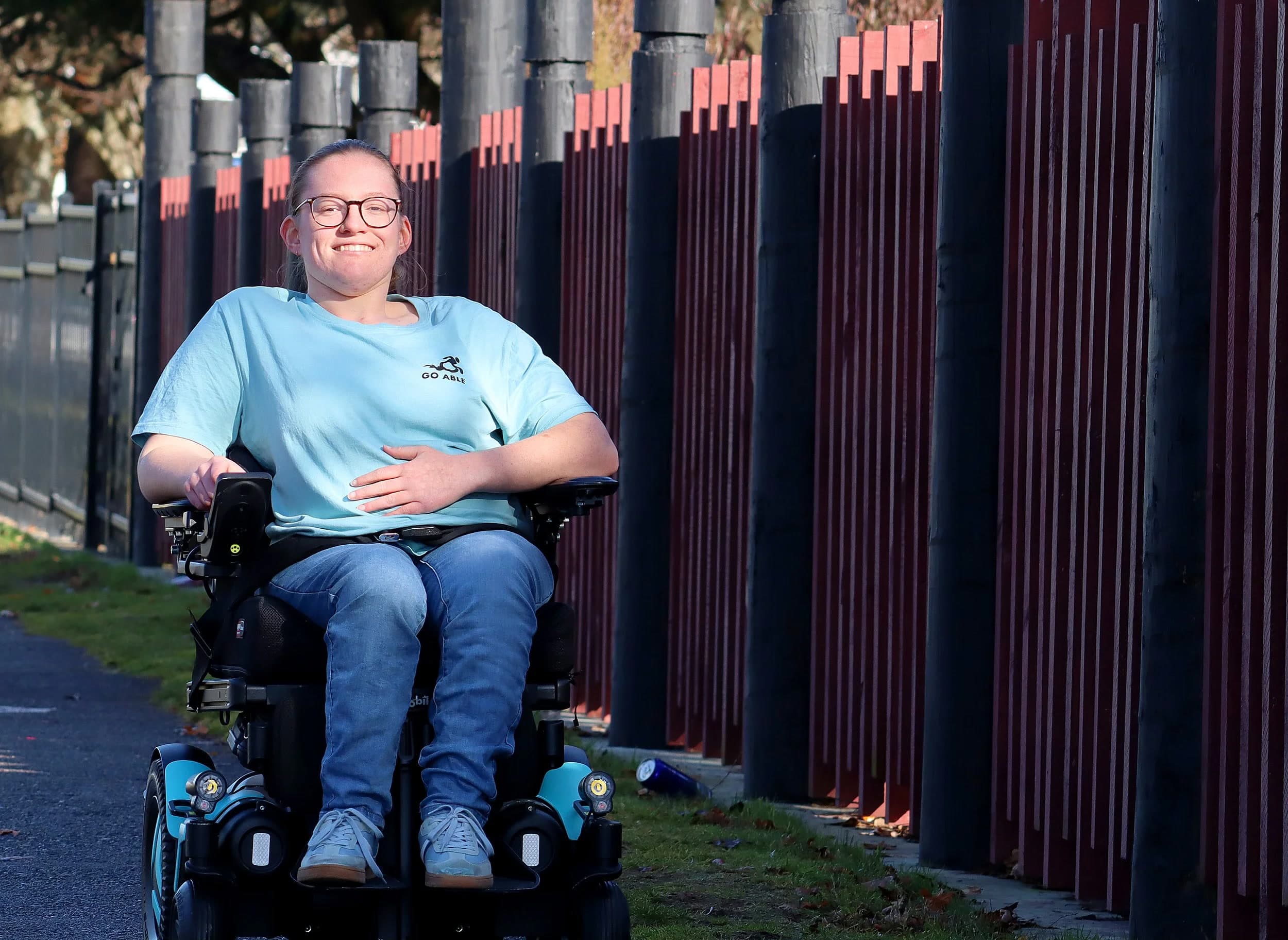 Daisy Hoe smiling in her wheelchair on a paved path, wearing a Go Able t-shirt