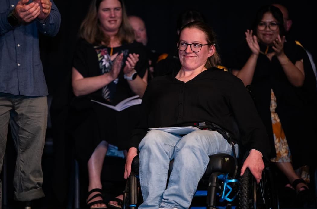 Daisy in her wheelchair at a national awards ceremony, smiling as the audience applauds